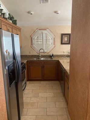 A kitchen with a stainless steel refrigerator, a built-in oven, a granite countertop, wooden cabinets, and a window with a geometric design.