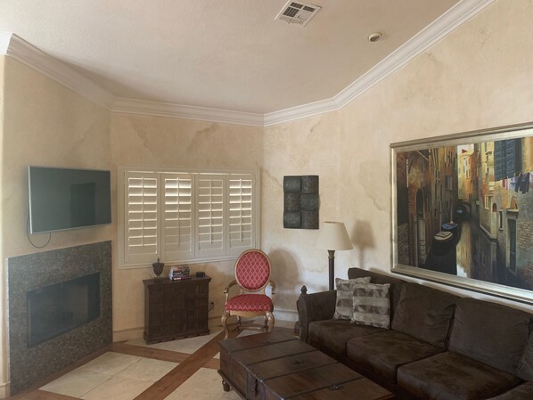 A living room with a television, a window with blinds, a chair, a sofa, a lamp, and a painting on the wall.