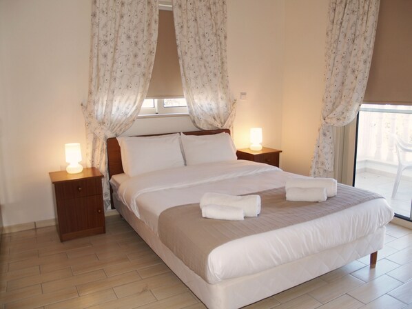 Una cama bien hecha con sábanas blancas y dos almohadas, con mesitas de noche de madera y lámparas, en una habitación con un ventanal y cortinas.