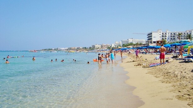 A beach with clear water, people swimming and sunbathing, and buildings in the background.