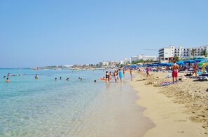 A beach with clear water, people swimming and sunbathing, and buildings in the background.