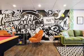 A room with a pool table, a green sofa, an orange chair, and a mural with text and graphics.