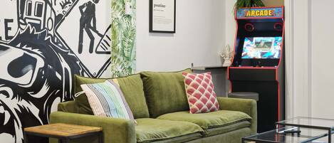 A living room with a green sofa, a small table, a framed picture, and a retro arcade machine.
