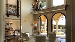A spacious living area with a high ceiling, a chandelier, and a view of a garden through large arched windows.