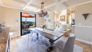 A dining area with a wooden table, clear chairs, and a chandelier. There is a view through a glass door to an outdoor patio.