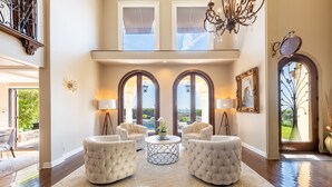 A spacious living room with a high ceiling, two large arched windows, a chandelier, and a round coffee table with a plant.