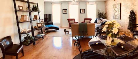 A cozy living room with a wooden floor, a glass coffee table, a red armchair, a blue sofa, and a bookshelf.