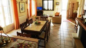 Un comedor con una mesa lista para cuatro, televisión y sofá en una habitación luminosa con ventanas grandes.