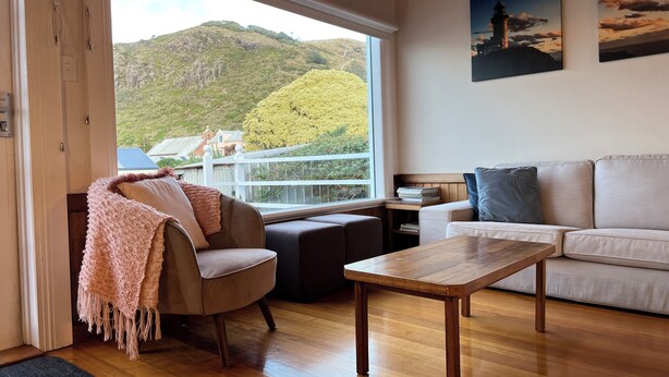 A modern living room with a wooden floor, a beige sofa, a wooden coffee table, and a chair with a pink blanket. A large window offers a view of a mountain and a house. - The Lodge of Stanley (Stanley)