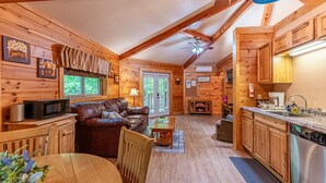 Interior acogedor de una cabaña de madera, con sala de estar, comedor, cocina y un ventanal grande.