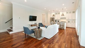 Uma sala de estar moderna com um sofá branco, poltronas azuis e uma mesa de centro de madeira. Uma televisão de tela plana está fixada na parede.