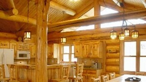 Interior rústico de una cabaña de madera con una gran mesa de comedor, varias sillas de madera y una zona de cocina con electrodomésticos básicos.