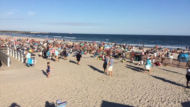 Ein belebter Strand mit Menschen, die laufen und sitzen, ein klarer Himmel und das Meer im Hintergrund.