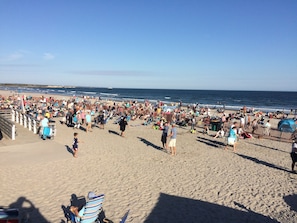 Una playa concurrida con gente caminando y sentada, un cielo despejado y el océano al fondo.