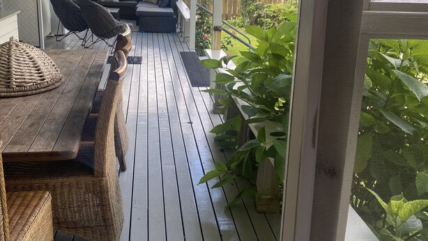En overdækket terrasse med træterrasse, fletmøbler og udsigt til baghaven.