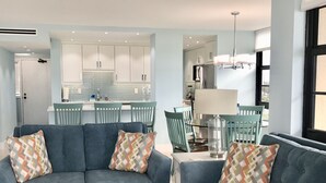 Ein modernes Wohnzimmer mit zwei blauen Sofas, einem Couchtisch und Kissen mit Muster. Im Hintergrund ist ein Essbereich mit Tisch und Stühlen zu sehen.