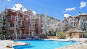Un espace piscine avec une eau d’un bleu limpide, entouré d’immeubles à plusieurs étages dotés de balcons, et un paysage montagneux en toile de fond.