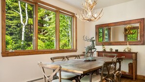 Une salle à manger avec une grande table en bois, six chaises, un lustre et une grande fenêtre donnant sur la forêt.
