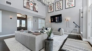 A spacious living room with a large gray sectional sofa, a wooden coffee table, and a decorative vase with flowers. There are framed artworks on the walls, a chandelier, and a mounted television. - Pool + Cabana + Amazing Views - Picturesque Point (Austin)