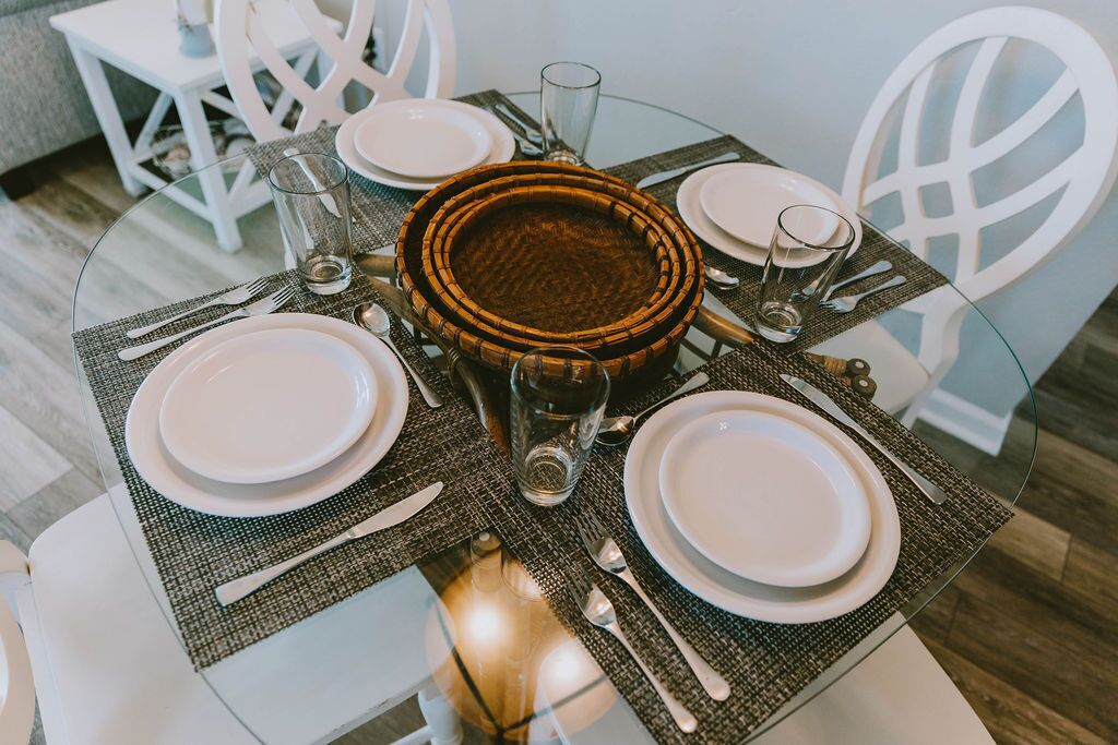 Une table de salle à manger ronde, avec quatre assiettes blanches, un saladier en osier au centre et un verre d’eau.