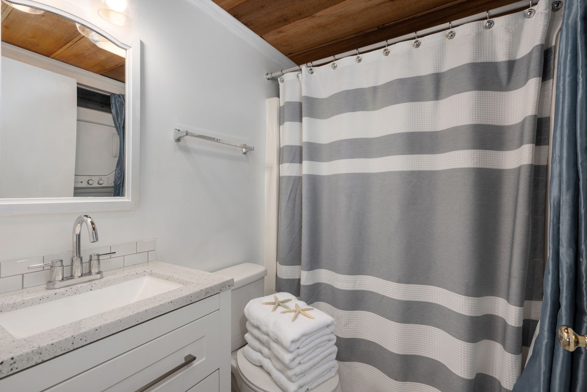 Un baño con lavamanos blanco, espejo, pila de toallas y cortina de ducha a rayas.