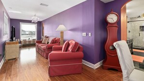 Una sala de estar con paredes moradas, piso de madera, un sofá rojo, una silla blanca, un reloj y una lámpara.