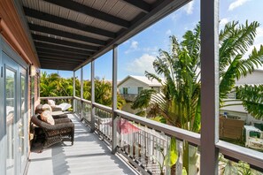 Un balcón techado con mobiliario de mimbre, barandilla blanca y vista a una zona residencial con palmeras y casas.