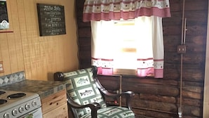 Interior rústico de una cabaña con un horno de leña, un sillón mecedor con cojines estampados con motivos de peces, una ventana con cortinas rosas y un letrero en la pared.