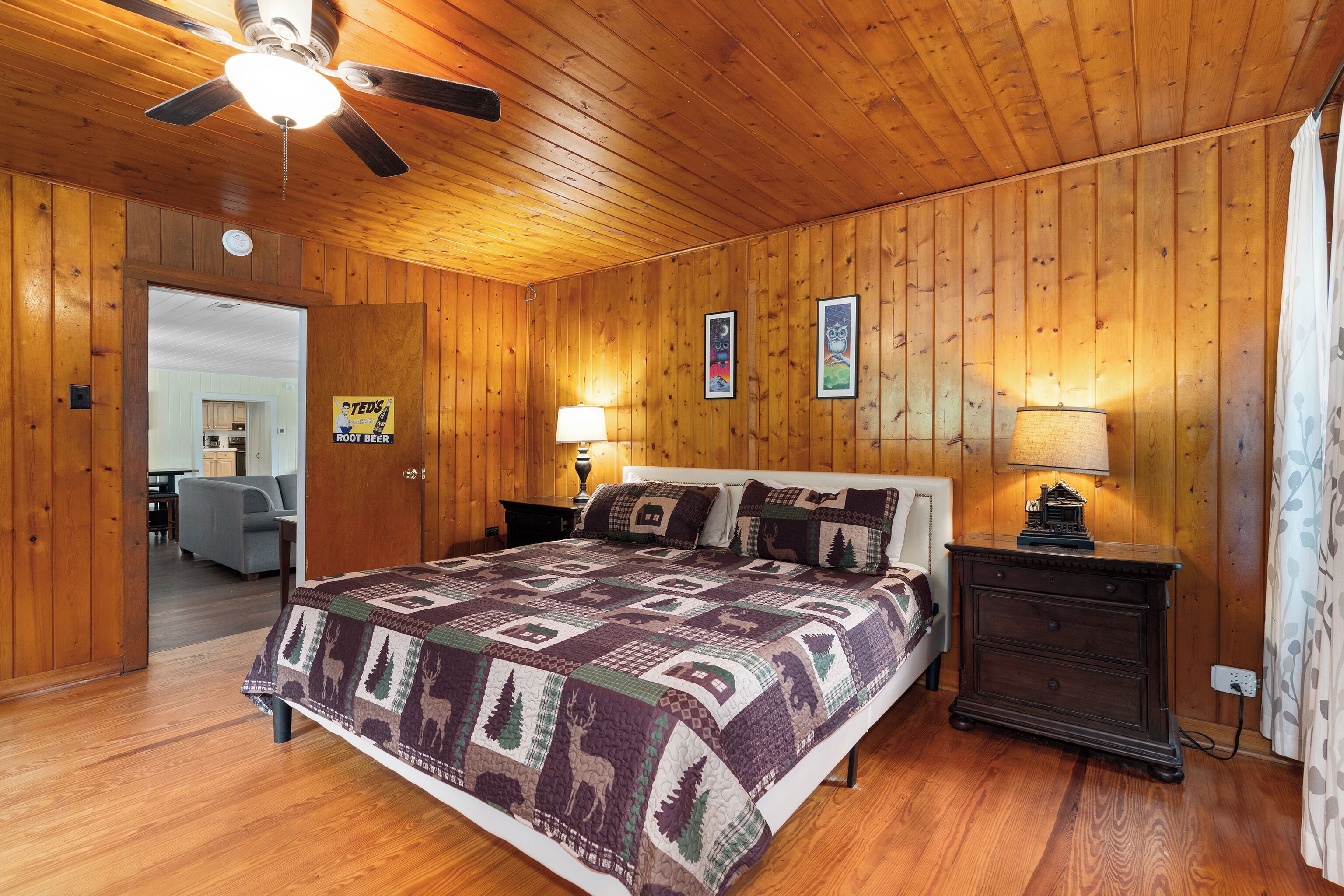 A wooden-paneled bedroom with a bed, two bedside tables with lamps, a ceiling fan, and a door leading to another room.