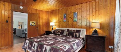 A wooden-paneled bedroom with a bed, two bedside tables with lamps, a ceiling fan, and a door leading to another room.