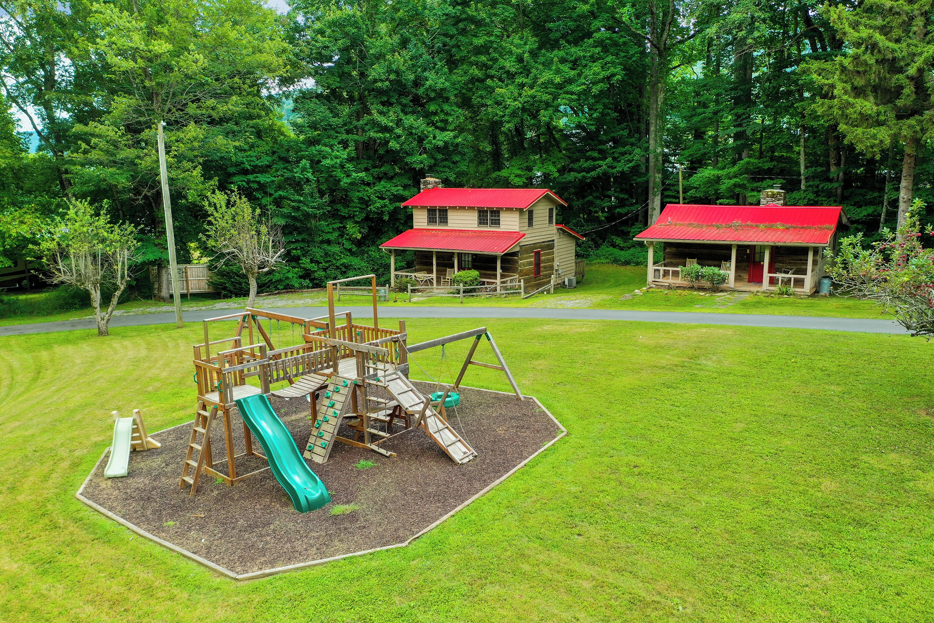 A playground with a slide and climbing structures, surrounded by a wooden cabin with a red roof.