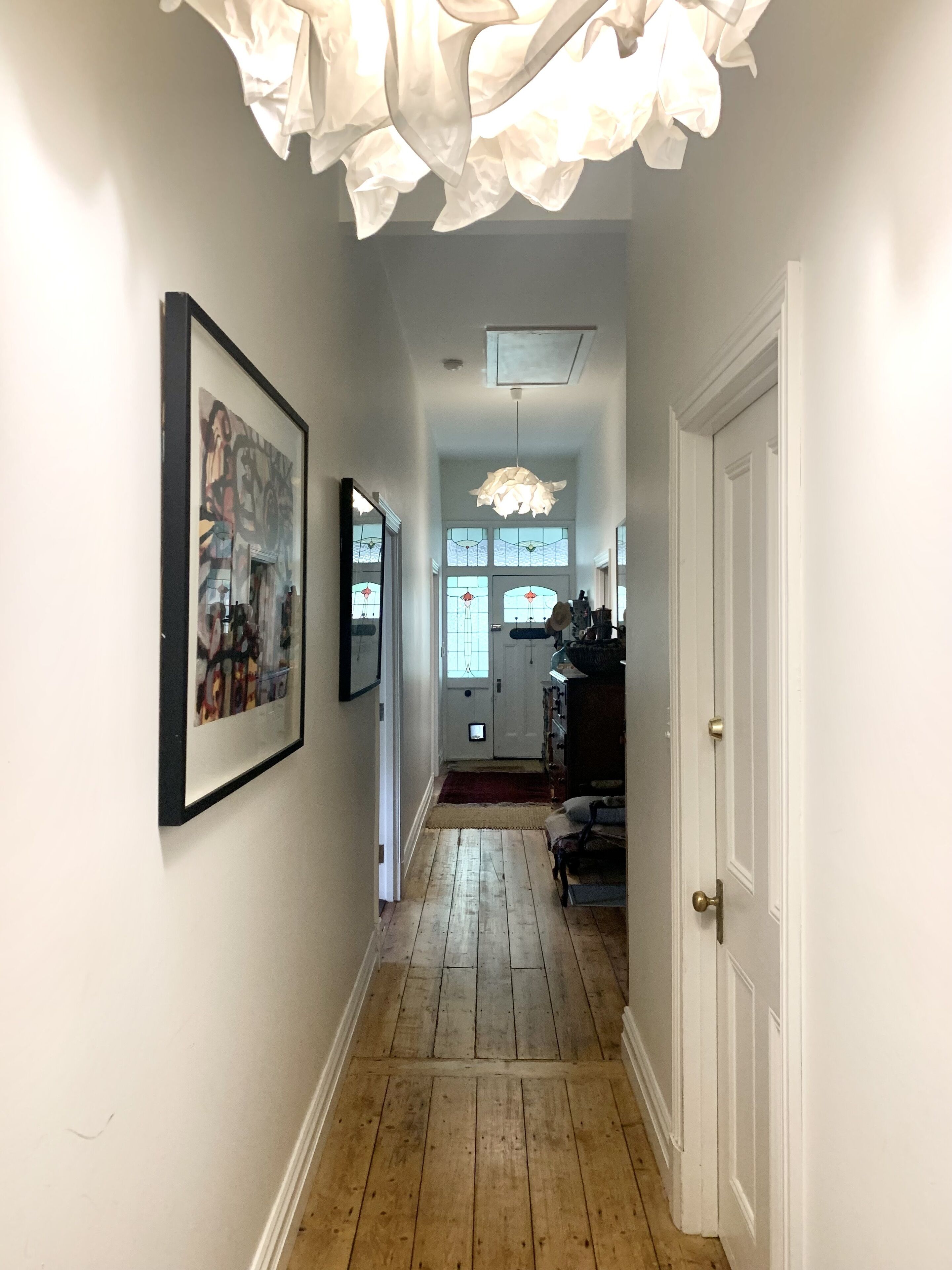 A hallway with wooden flooring, white walls, and a ceiling light fixture. There are framed artworks on the walls and a door on the right.