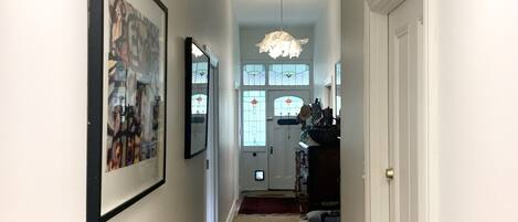 A hallway with wooden flooring, white walls, and a ceiling light fixture. There are framed artworks on the walls and a door on the right.