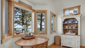 Uma área de refeições com mesa e cadeiras de madeira, um armário branco com louças azuis e brancas, e grandes janelas com vista para árvores e um corpo d'água.