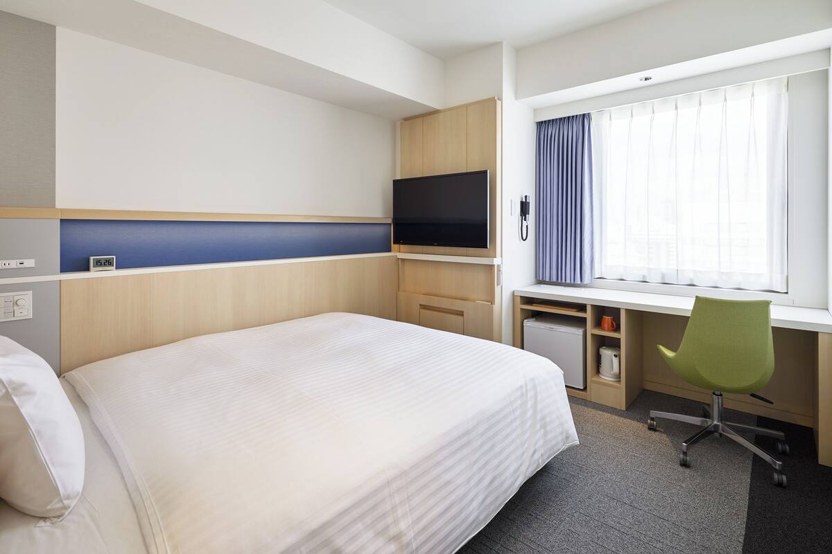 A hotel room with a large bed, a desk with a computer and chair, a TV, and a window with curtains.