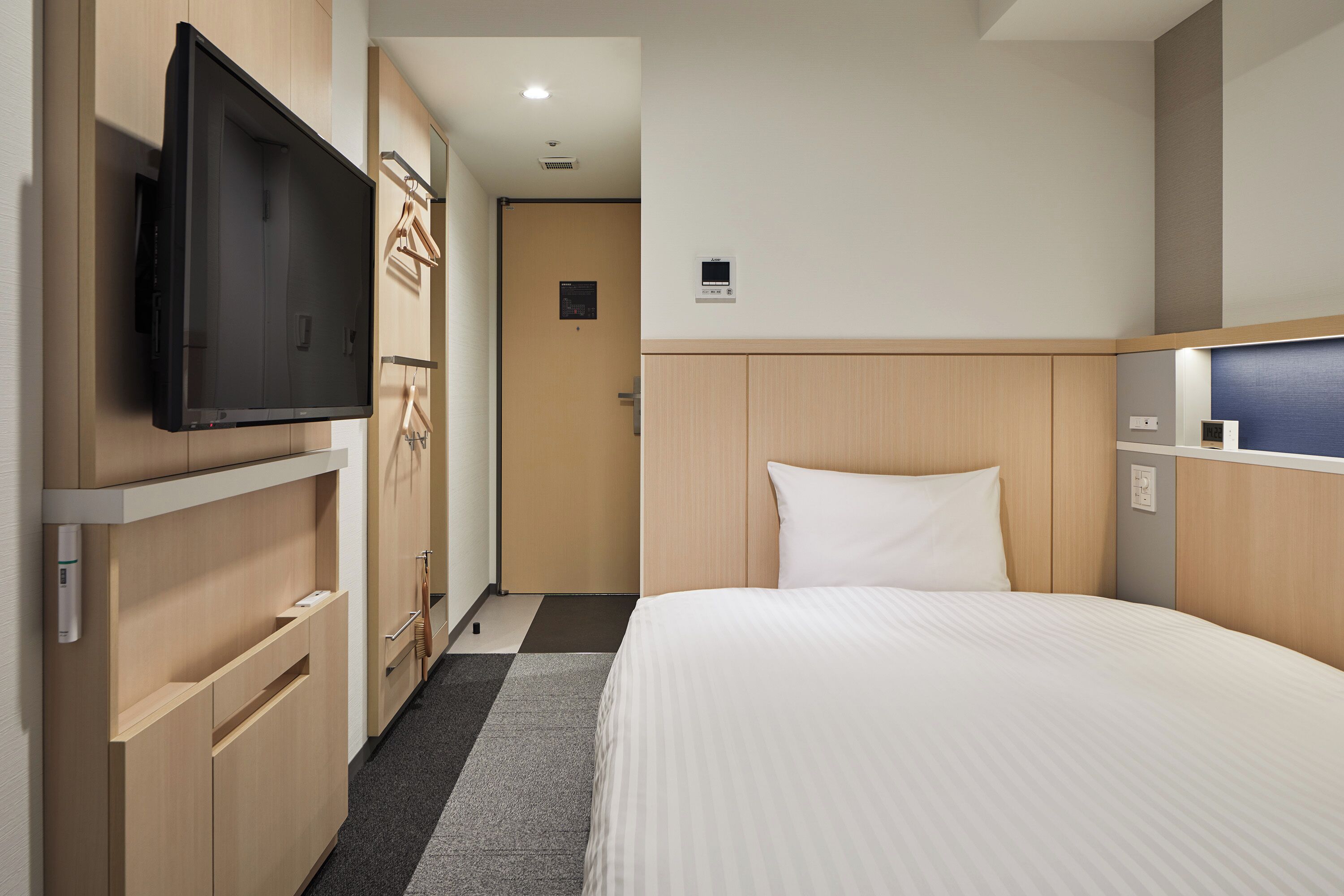 A hotel room with a bed, a television mounted on the wall, a wardrobe, and a small desk with a mirror.