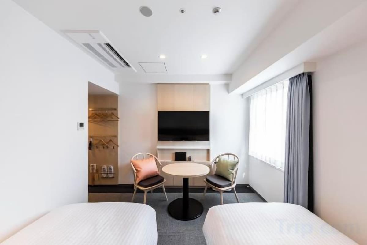Une chambre d’hôtel avec deux lits, une télévision, une petite table ronde avec des chaises et une fenêtre avec des rideaux.