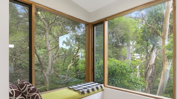 A room with large windows, a green cushioned bench, a small round table with a book, and a decorative pillow. - Clovelly (Lorne)