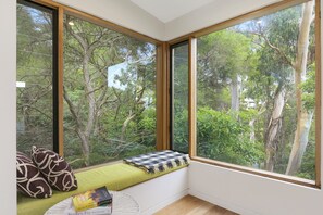 A room with large windows, a green cushioned bench, a small round table with a book, and a decorative pillow.