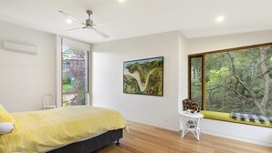 A bedroom with a bed, a ceiling fan, a window with a view of trees, and a small table with books.