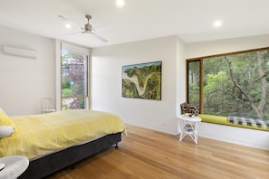 A bedroom with a bed, a ceiling fan, a window with a view of trees, and a small table with books.