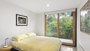 A bedroom with a bed, a bedside table with a lamp, a framed picture on the wall, and a sliding glass door leading to an outdoor area. - Clovelly (Lorne)