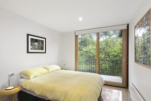 A bedroom with a bed, a bedside table with a lamp, a framed picture on the wall, and a sliding glass door leading to an outdoor area. - Clovelly (Lorne)