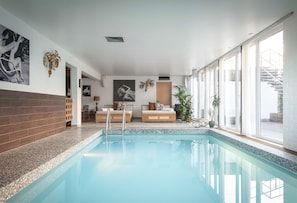 Una piscina interior con borde de azulejos, un banco de madera y un sofá con cojines. Hay dos cuadros enmarcados en la pared, una planta en maceta y una ventana con vistas al exterior.