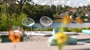 Una terraza con dos sillas blancas de metal, una mesa con una copa de vino y una vista de un lago y casas al fondo.