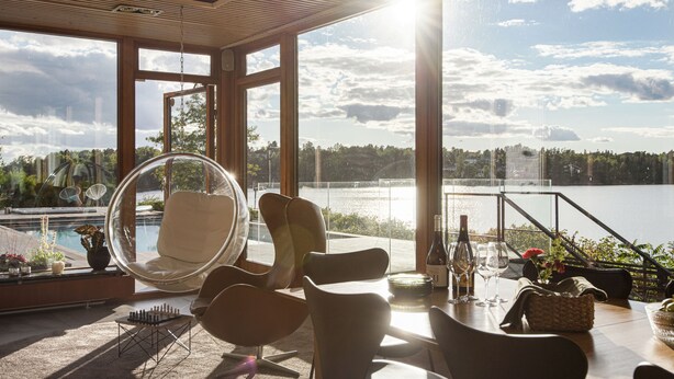 Un área de estar moderna al aire libre con vista a un lago, que incluye una silla redonda, sillones de descanso y una mesa de comedor con copas y botellas de vino.