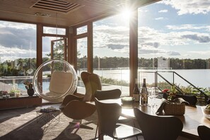 Un área de estar moderna al aire libre con vista a un lago, que incluye una silla redonda, sillones de descanso y una mesa de comedor con copas y botellas de vino.