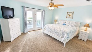Une chambre à coucher avec un lit, un téléviseur posé sur une commode, un ventilateur de plafond et un balcon avec des rideaux.