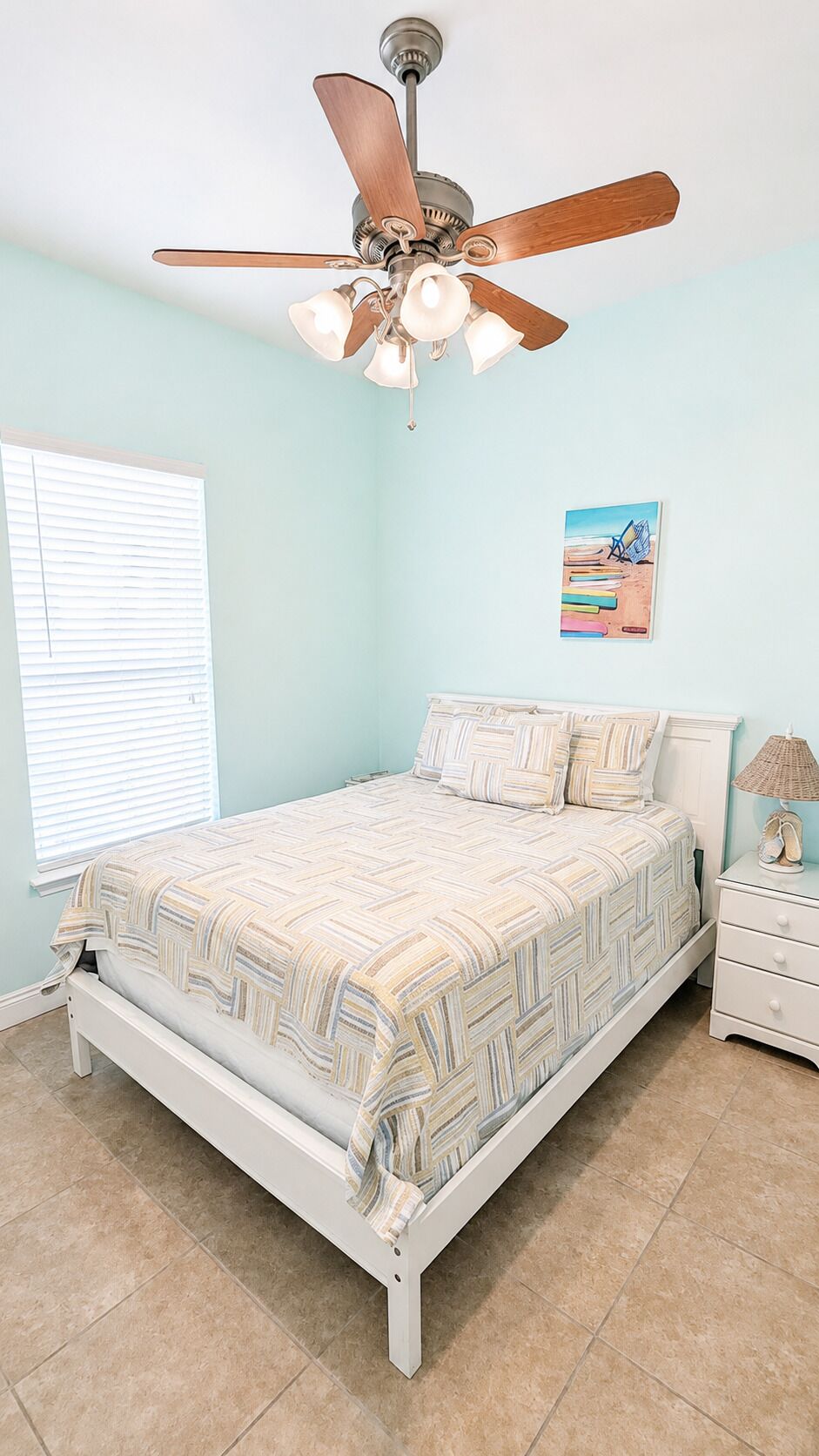 A bedroom with a bed, a nightstand, a lamp, a ceiling fan, and a beach-themed painting on the wall.