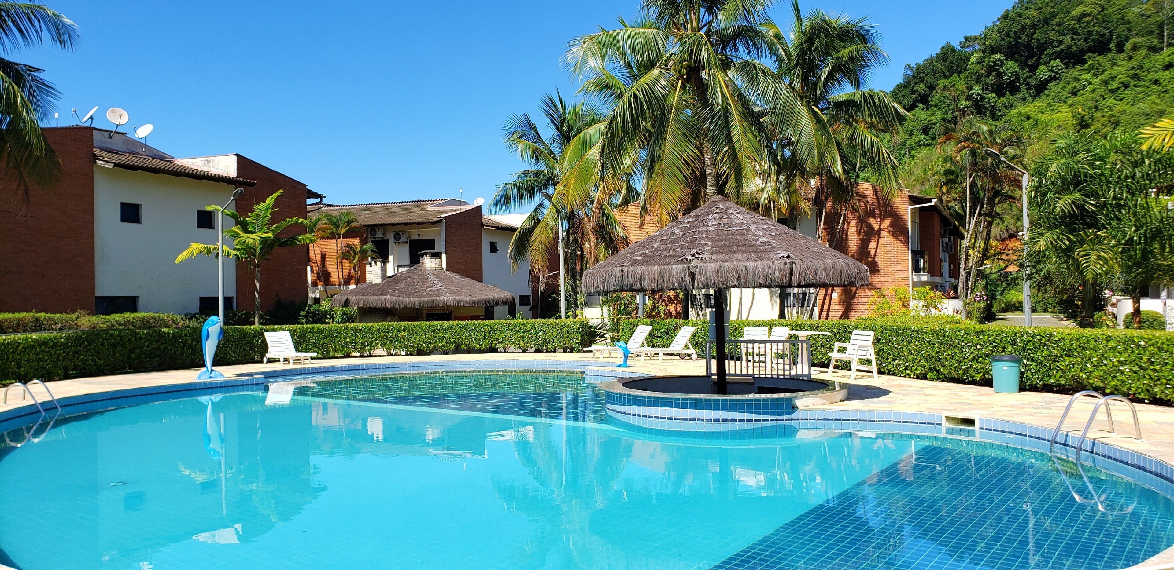 A swimming pool with lounge chairs and thatched umbrellas, surrounded by palm trees and a building with satellite dishes.